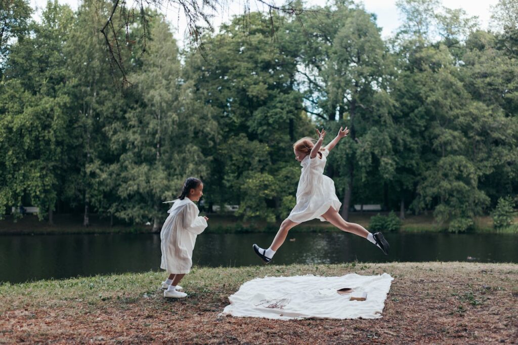 pexels photo 10653640 Two girls in white dresses playing joyfully by a lake in a lush green park.