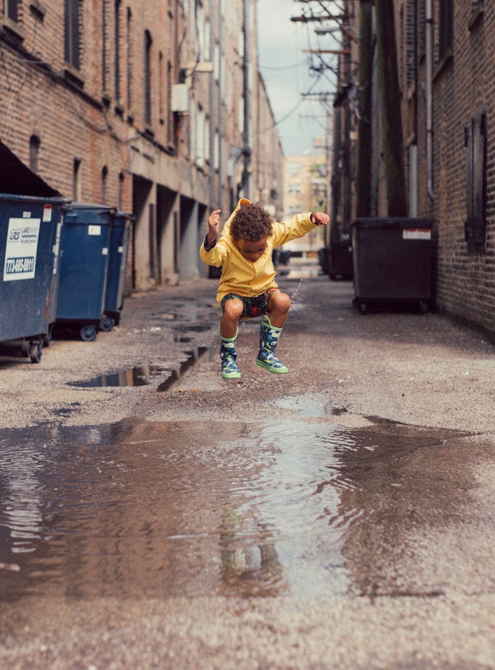 pexels photo 2387702 A young boy in a yellow outfit jumps over a puddle in an urban alley. Vibrant and playful moment.